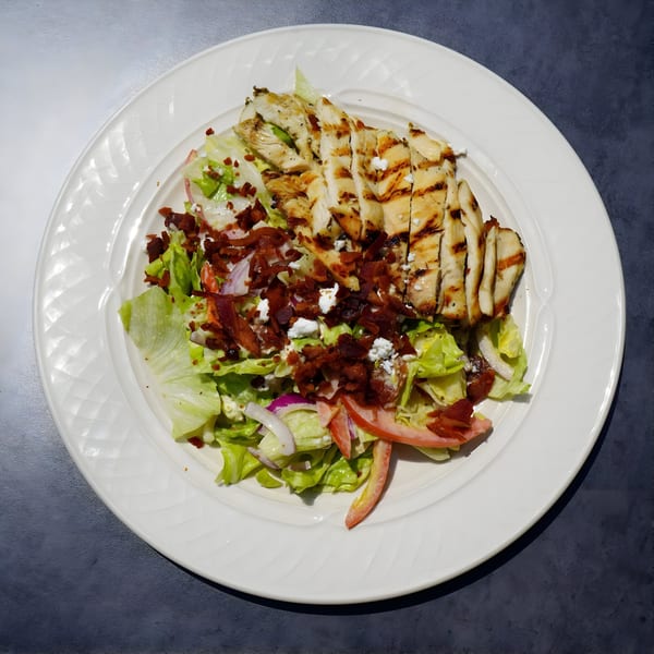 Grilled chicken salad with lettuce, bacon, feta, red onion, and tomato on a white plate.