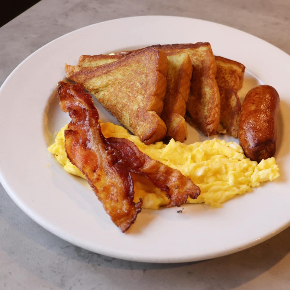 A diner breakfast plate with French toast, scrambled eggs, crispy bacon, and a sausage link.