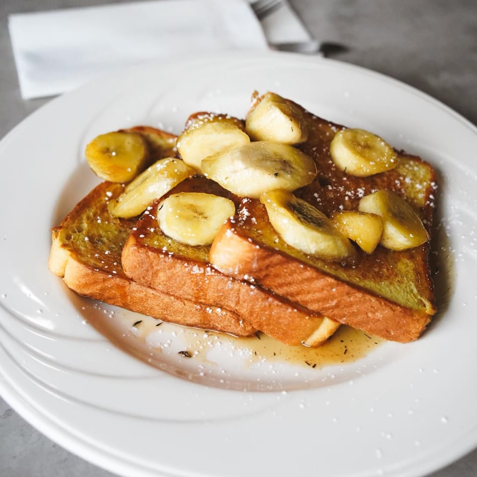 Golden French toast slices topped with caramelized bananas, powdered sugar, and syrup on a white plate.