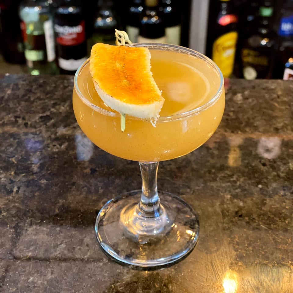 A golden cocktail in a coupe glass, garnished with a caramelized banana slice, on a dark bar.