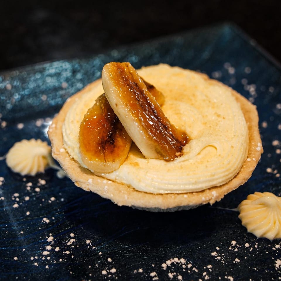 Banana cream pie tart with caramelized bananas and powdered sugar on a dark blue plate.
